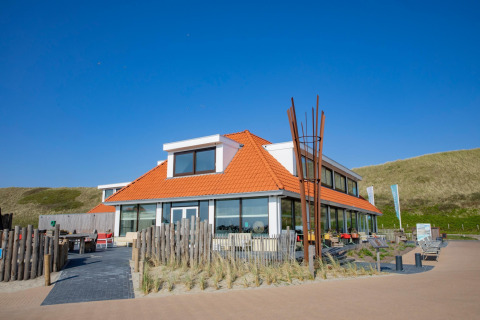 Foto van een eigentijdse lodge met oranje dak, terras, blauwe lucht en omliggende duinen.