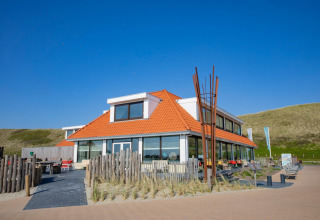 Foto van een eigentijdse lodge met oranje dak, terras, blauwe lucht en omliggende duinen.