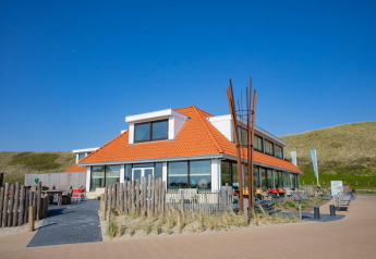 Foto af en moderne lodge med orange tag ved klitterne, blå himmel og udendørs siddepladser.
