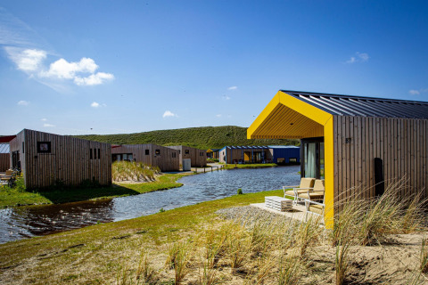 Moderne houten lodges met gele randen aan een vijver, omgeven door gras, duinen en blauwe lucht.