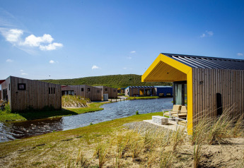 Moderne trælodges med gule detaljer ved en sø, omgivet af græs og bakker under en klar blå himmel.