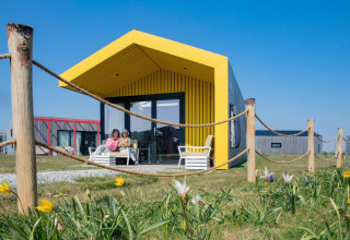 Two people sit on the porch of a modern lodge with a yellow roof, surrounded by a rope fence and flowers.