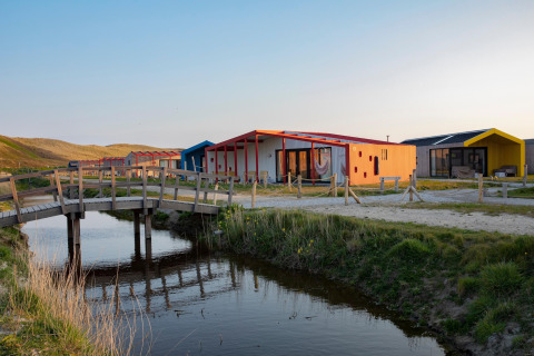 Colorful lodge cabins by a stream, with a wooden bridge and grassy dunes in the soft evening sunlight.