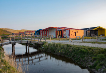 Des lodges colorés en bord de ruisseau, avec un pont en bois et des dunes herbeuses au coucher du soleil.