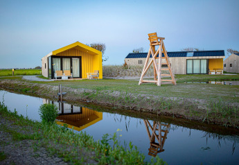 Photo de lodges modernes près d’un canal au coucher du soleil avec reflets et accents jaunes et bois.