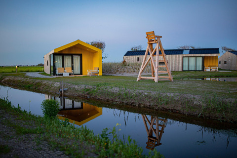 Photo of modern lodge cabins by a canal at sunset with yellow accents, wood features, and reflections.