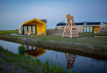 Billede af moderne lodgehytter ved en kanal med gult og træ, spejlet i vandet, ved solnedgang.