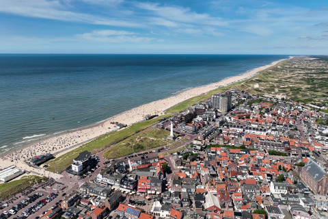 Luchtbeeld van een kuststad met zandstrand, zee en rode daken onder een stralend blauwe hemel.