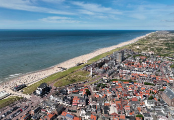 Luftfoto af en kystby med strand, hav og røde tage under en klar blå himmel på en solrig dag.
