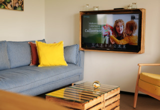 Living room at Resort Callantsoog 1 with blue sofa, yellow pillow, wall TV, and glass coffee table in the Netherlands.