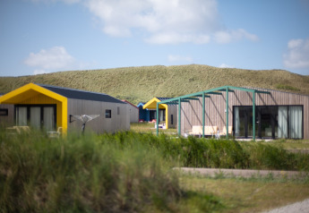 Lodges modernes aux toits jaunes devant des dunes herbeuses sous un ciel bleu, ambiance de vacances.
