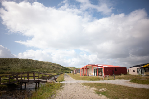 Pathway to Resort Callantsoog 1 in the Netherlands, with a wooden bridge, cabins, dunes, and a cloudy sky.
