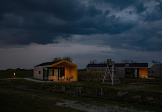 Avondbeeld van sfeervolle houten lodges met warme verlichting, op een groene weide onder donkere wolkenlucht.