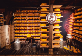 Estantes llenos de ruedas de queso en un almacén con un cartel de Henri Willig y latas de leche.