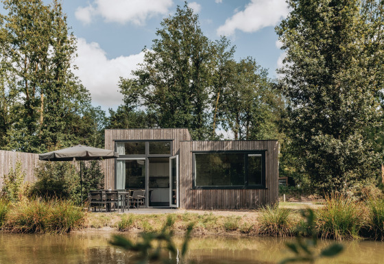 Tiny house Holiday Park Schaijk 3, modern houten huisje aan een vijver, omringd door bomen en natuur.