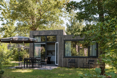 Modern tiny house at Holiday Park Schaijk with wood exterior, large windows, and outdoor seating in greenery.