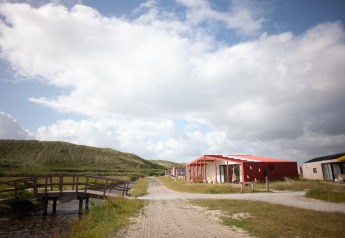 Vue du Resort Callantsoog 5 aux Pays-Bas, avec chalets rouges, pont en bois et dunes sous un ciel nuageux.