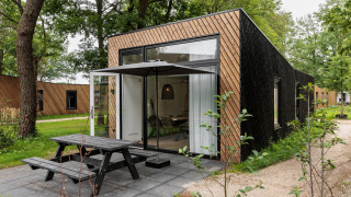 Moderne tiny house med store vinduer, terrasse og bænk i skovomgivelser, ideel til afslapning.