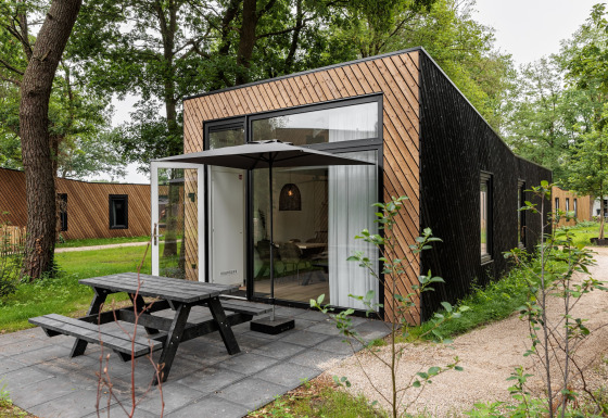 Moderne tiny house med store vinduer, terrasse og bænk i skovomgivelser, ideel til afslapning.