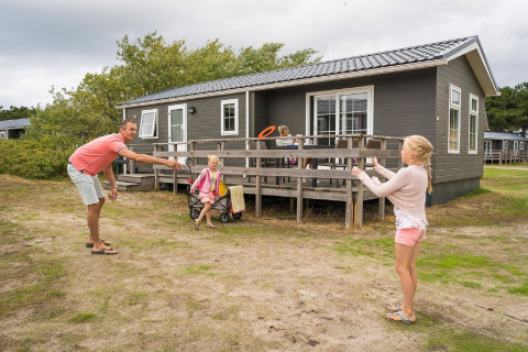 Familie spielt vor einer Holzhütte im Holiday Park Loodsmansduin 1 in den Niederlanden bei bewölktem Himmel.