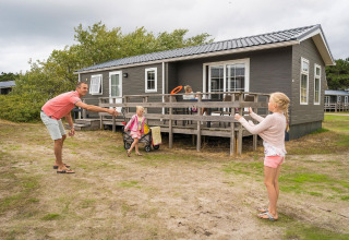 Familie spielt vor einer Holzhütte im Holiday Park Loodsmansduin 1 in den Niederlanden bei bewölktem Himmel.