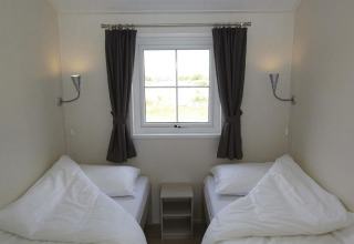 Deux lits simples dans une chambre claire, fenêtre centrale, rideaux sombres et table de chevet.