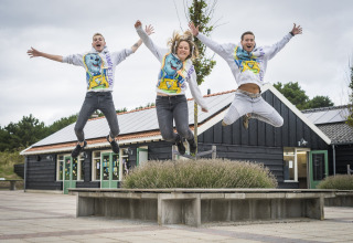 Tre unge mennesker hopper glade foran Holiday Park Loodsmansduin 1, en lodge i Holland med sorte bygninger.