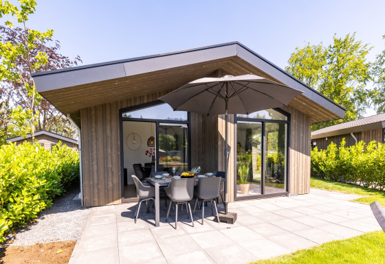 Moderne chalet i Holiday Park Molenvelden 8 med terrasse, havemøbler og stor parasol omgivet af grønt.