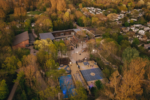 Vista aérea de una cabaña rodeada de árboles, canchas deportivas y varias construcciones al atardecer.