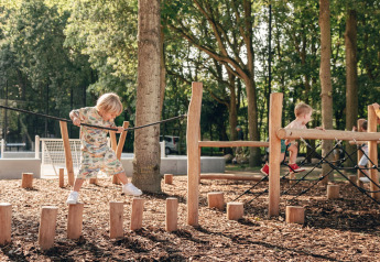 Kinder spielen auf einem Outdoor-Spielplatz mit Holzpfosten und Klettergerüst in einer bewaldeten Lodge.