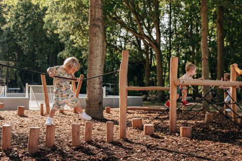 Niños juegan en un parque infantil al aire libre con postes de madera en un entorno de cabaña boscosa.