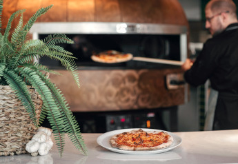 Ein Koch backt Pizza im Steinofen in einer Lodge, mit Farnpflanze und Knoblauch auf dem Tresen.