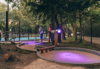 Persone giocano a minigolf al crepuscolo vicino a un lodge, con luci viola che illuminano la foresta.