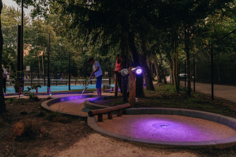 People play mini-golf at dusk by a lodge, with purple lighting illuminating the course in a forest setting.