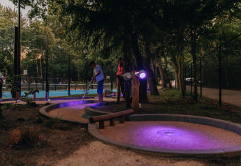 People play mini-golf at dusk by a lodge, with purple lighting illuminating the course in a forest setting.