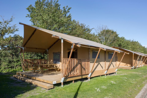 Safari tent with a wooden porch and picnic table, set on a grassy area with trees under clear blue sky.