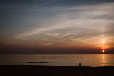 Due persone passeggiano sulla spiaggia al tramonto vicino a un lodge, sotto un cielo sereno e colorato.