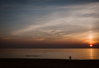 Due persone passeggiano sulla spiaggia al tramonto vicino a un lodge, sotto un cielo sereno e colorato.