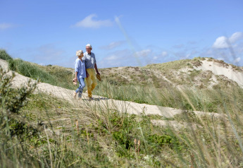 Älteres Paar spaziert barfuß auf einem Dünenweg, umgeben von Gras, unter blauem Himmel.