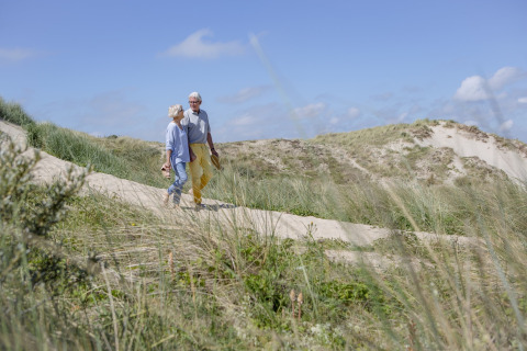 Coppia anziana cammina a piedi nudi su un sentiero tra le dune, circondata da erba e cielo azzurro.