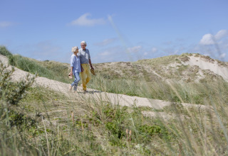 Coppia anziana cammina a piedi nudi su un sentiero tra le dune, circondata da erba e cielo azzurro.
