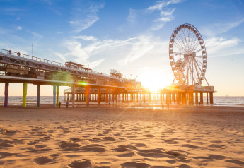 Sunset view of a pier with a Ferris wheel and people walking on the sandy beach by the sea.