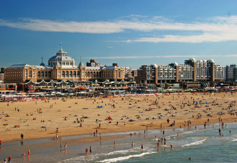 Scena di spiaggia affollata davanti a un hotel storico e a edifici moderni sotto un cielo sereno.