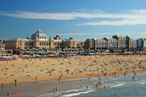 Beach scene with many people relaxing in front of a historic hotel and modern buildings on a sunny day.