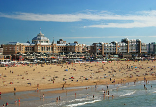 Strandscene med mange mennesker foran et historisk hotel og moderne bygninger under blå himmel.