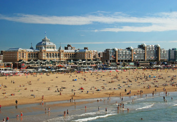 Beach scene with many people relaxing in front of a historic hotel and modern buildings on a sunny day.