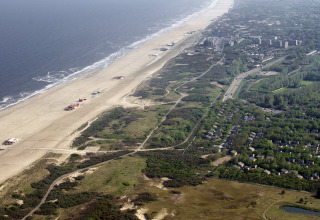 Vista aerea di un lodge sulla spiaggia con bungalow, dune, una strada e l’oceano che si estende lontano.