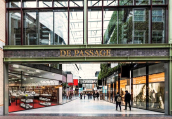 Eingang zur Einkaufspassage 'De Passage' mit Glasdach, Geschäften und Passanten in moderner Architektur.