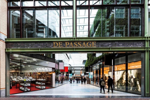 Indgangen til De Passage indkøbscenter med glasloft, butikker og mennesker, der går forbi.