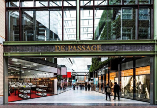 Toegang tot winkelgalerij De Passage met glazen dak, winkels en voorbijgangers in de overdekte passage.